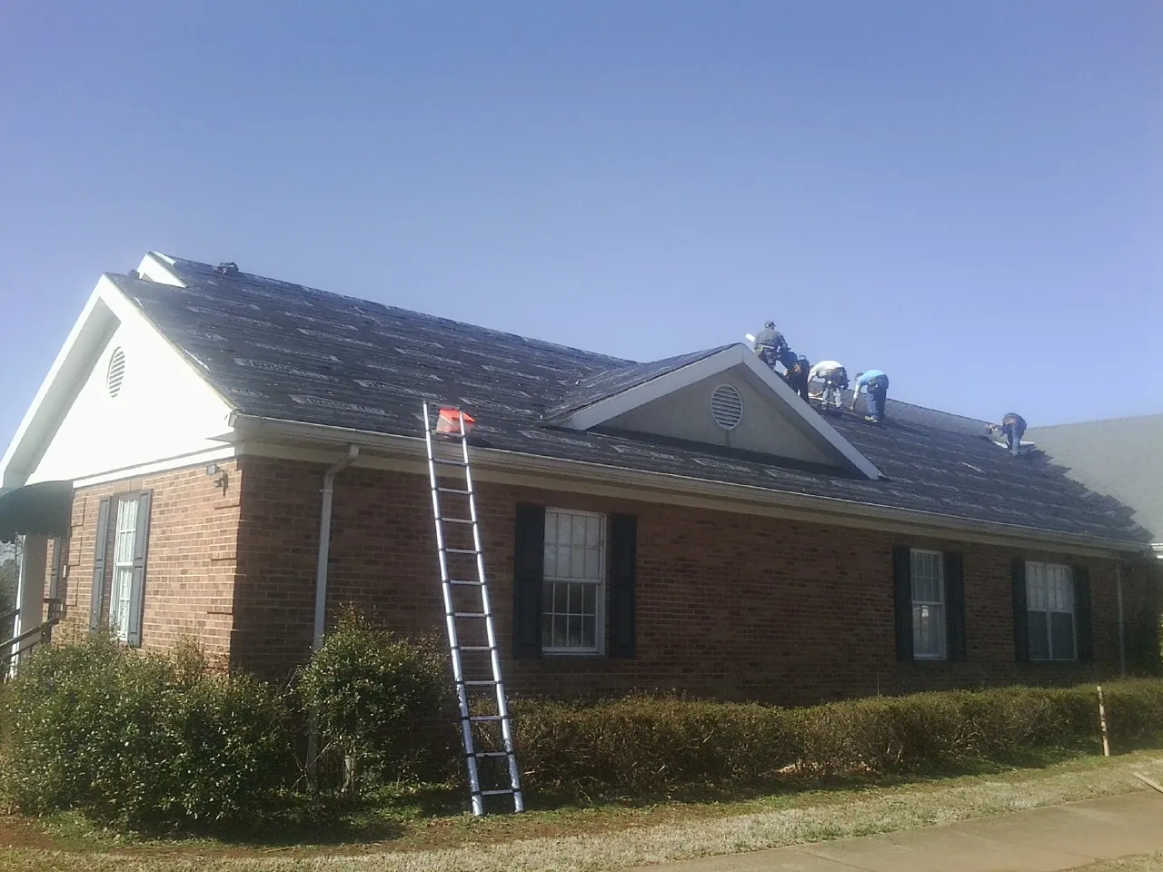 Crew tearing shingles off a brick ranch roof with a ladder set — Metal Roof Repair work by our crew in Camilla