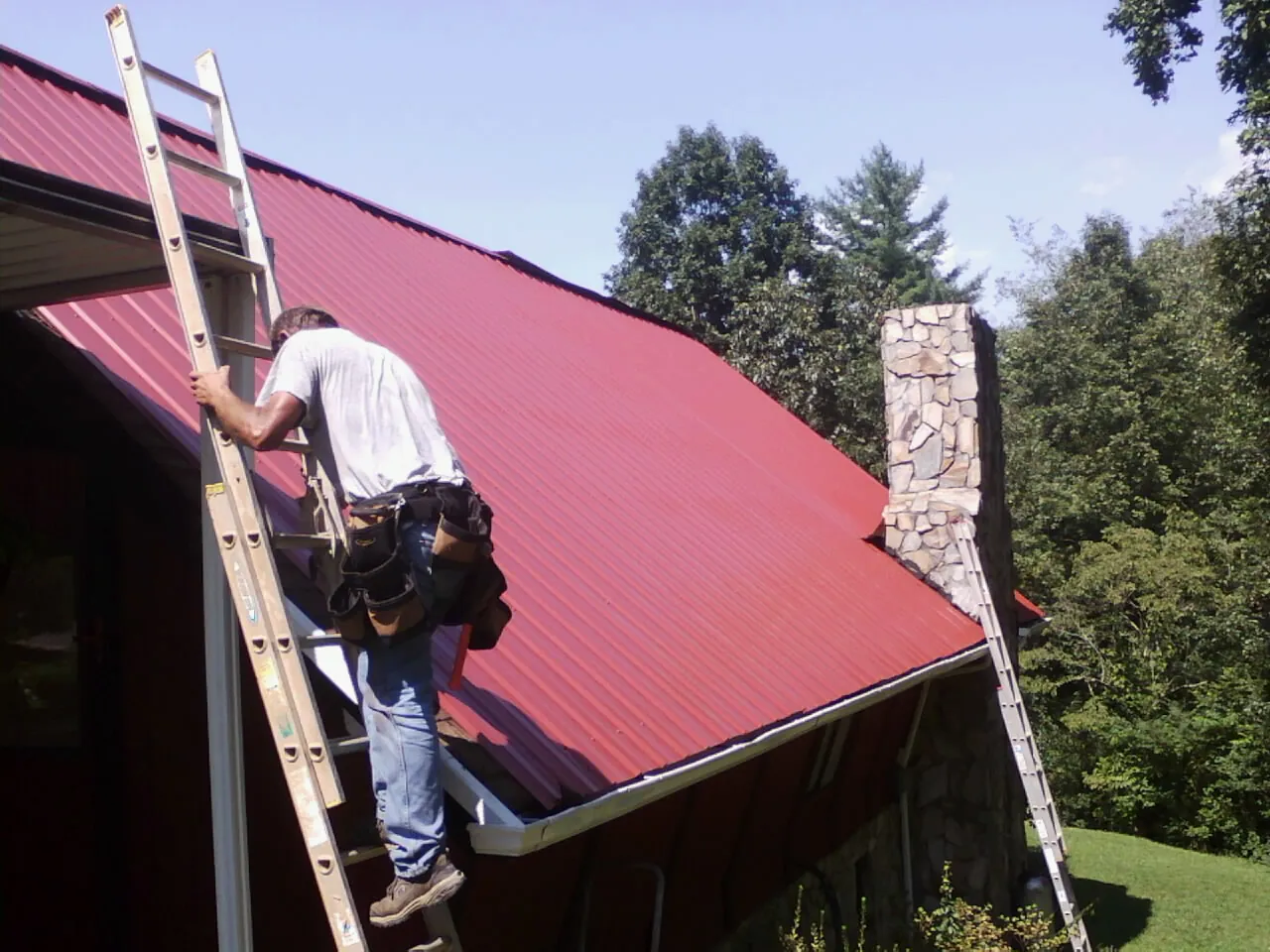 Roofer on a ladder beside a red metal roof and stone chimney — Soffit & Fascia Repair by Roofing Brothers in Camilla